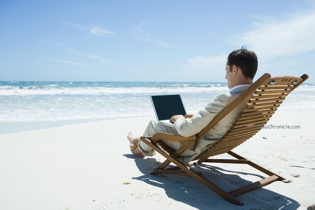 Online Booking - Businessman sitting in lounge chair on beach, barefoot, using laptop